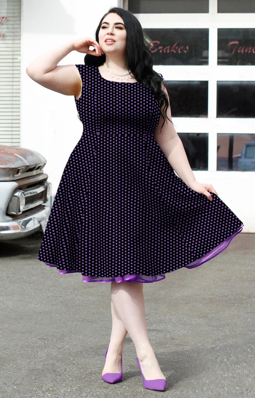 A curvy woman with long dark hair poses in a retro styled purple polka dot dress with a purple crinoline peeking from the hem it has a cinched waist and a square neckline and is gorgeous fun looking