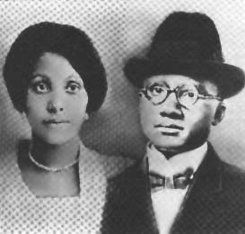 Louise as a young woman with her hair back and a bead necklace, next to her husband Earl who has glasses and a black hat and coat.