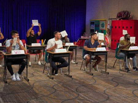 The guys sit at school desks and hold their answers up on white boards