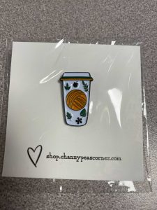 An enamel pin of an orange ball of yarn on a takeaway coffee cup