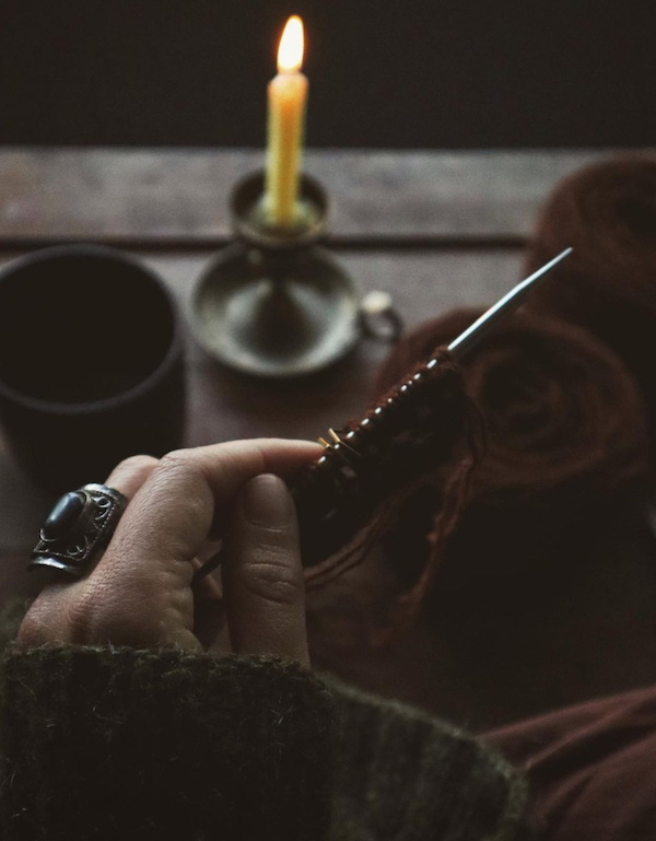 A dark moody shot of a White hand holding a knitting needle with stitches cast on in dark ruby red yarn with acandle burning in the background