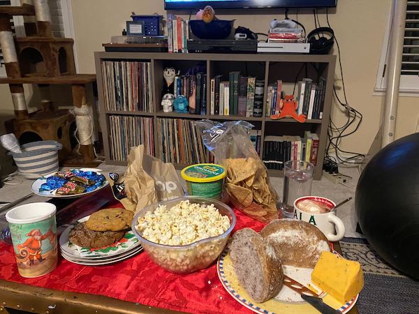 A coffee table covered with snack food like popcorn, cookies, twix bars, cheese, bread, crackers, dip and chips and guacamole Crap now I am hungry