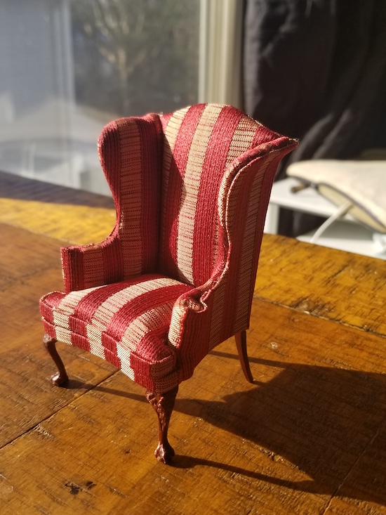 A miniature dollhouse wing chair with carved wooden legs and gold and red striped fabric
