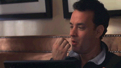 Tom Hanks sits upright in bed with a laptop on his lap. He's wearing an excited smile. He blows on his hands, then stabs one finger downward towards the keypad.