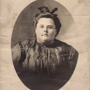 A sepia-toned photo of a white woman with a round face and small glasses, wearing a black formal Victorian dress