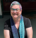 A photo of catherine with blue and pink short hair wearing a blue and pink crochet scarf she made herself she is smiling at the camera