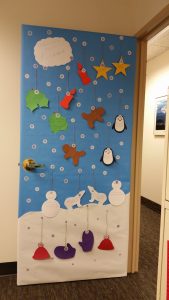Maggie's awardwinning door with a blue background and a white hill of snow at the bottomw with cutouts of penguins, snow hats and gingerbread men hanging from the door each with a pun joke written on them! 