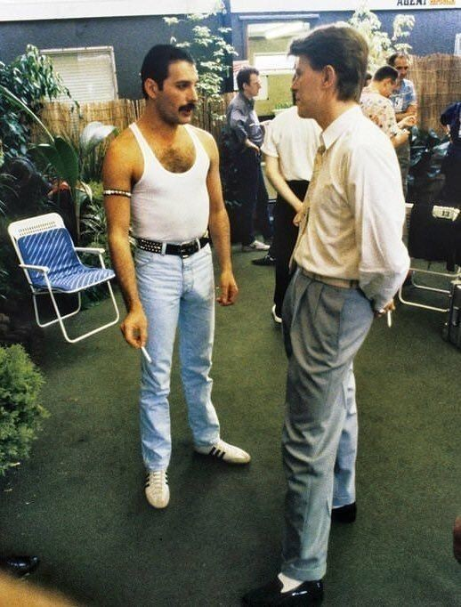 A photo of Freddie Mercury and David Bowie standing and talking, but their pants are very tight.