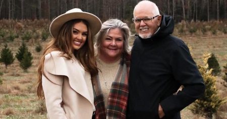 Susie standing with her parents. Shes' wearing a giant hat her hair is sort of in her face and she's wearing adrapey cream coat. Her mom has grey shoulder length hair, and her dad is balding, with a white beard and glasses.