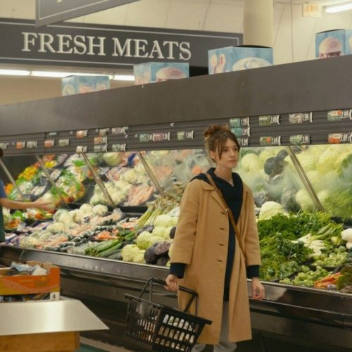 Noa doing her grocery shopping in a brown coat and sweatpants.