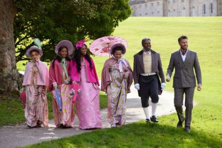 The court walks to the country fair. The women are in various shades of pink with hats and caps and two men are with them one in shorter pants and white gloves the other in a double breasted suit jacket and cravat 