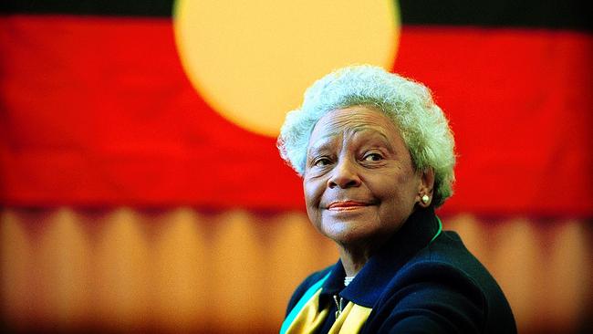 Colorful photo of Faith Bandler, with white hair and smiling in front of the Aboriginal Australian flag which is red and black with a yellow circle in the middle