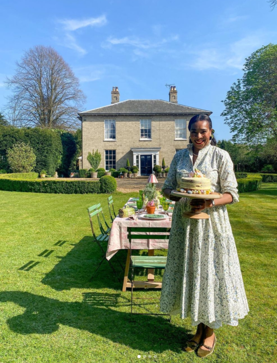 Paula Sutton of Hill House Vintage in front of her home with. table al fresco set for eight with green chairs. Paula is a Black woman with a beautiful smile and she is wearing red lipstick and a green print dress in this image, and she's holding a BIG CAKE 