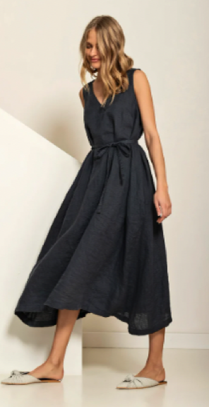 A blonde model wearing a linen long tank dress in dark grey with a waist tie and white slides the dress has pockets and a rather voluminous skirt