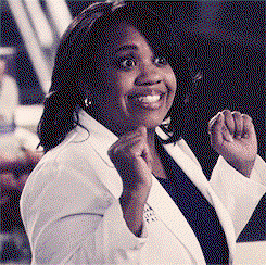 Chandra Wilson in a medical coat puts her arms above her head with a big smile 