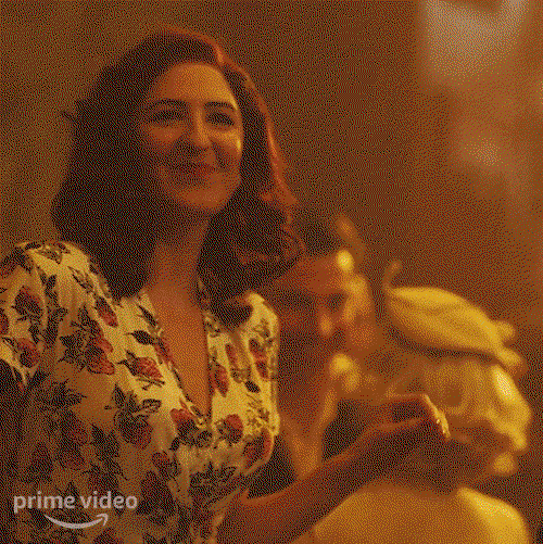 Greta, played by D'Arcy Carden, with shoulder length hair and a strawberry print blouse with a deep v neck waves and smiles at someone at a dance 