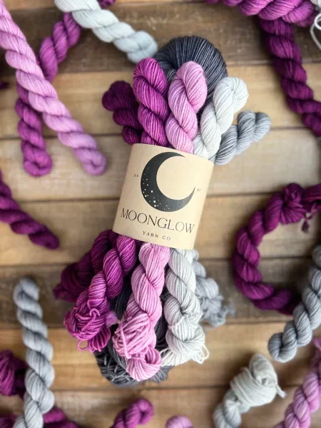 A skein of dark gray yarn with mini skeins in purple, pink, white and light gray