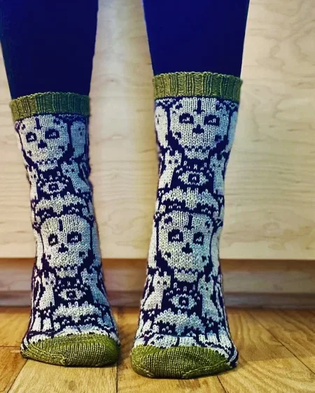 A pair of socks with a sour green cuff and toe. The body of the sock is purple with white skulls, cats and other spooky images