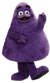 A still image of Grimace, who is a purple gumdrop shaped furry creature with legs and short arms. 