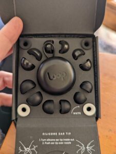 A picture of the box of Loops with a black circle carrying case in the center, and assorted foam and rubber tips plus mute options in each corner