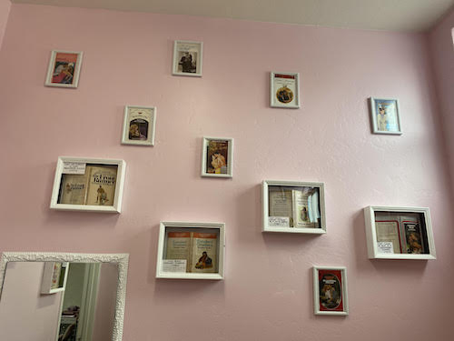 A pink wall with a high ceiling and white windowbox frames of some classic category romances and covers some open books and some just the front cover
