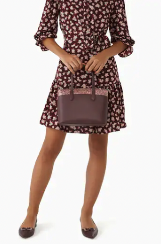 A photograph of a model holding the bag wearing a complimentary dress in burgundy floral print with burgundy shoes 