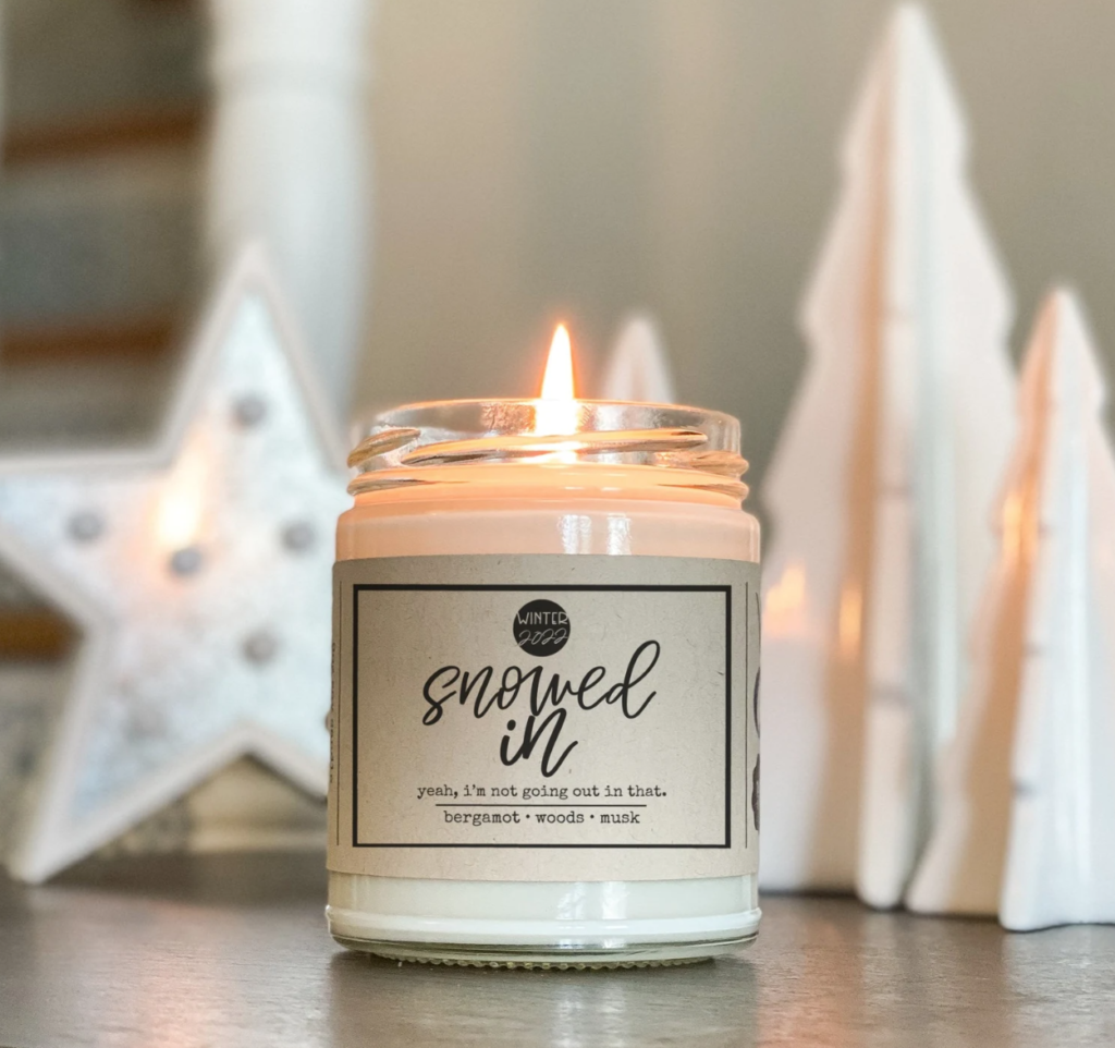 A to Z Candles Snowed In. A lit white candle in a glass jar with white star and tree decorations blurred in the background.