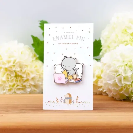 An enamel pin of a gray cat knitting in bed