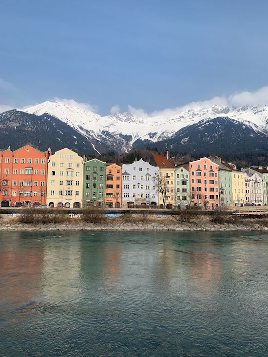 Pastel buildings reflected in the water of a lake with snow covered mountains in the background