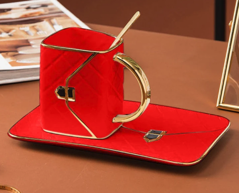 A red porcelain mug shaped like a red handbag with gold detail and handles, on top of a rectangular saucer that looks like a bag, with a gold spoon