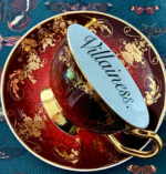 A deep red teacup and saucer with gold filigree accents and the word VILLAINESS written on the inside edge of the teacup