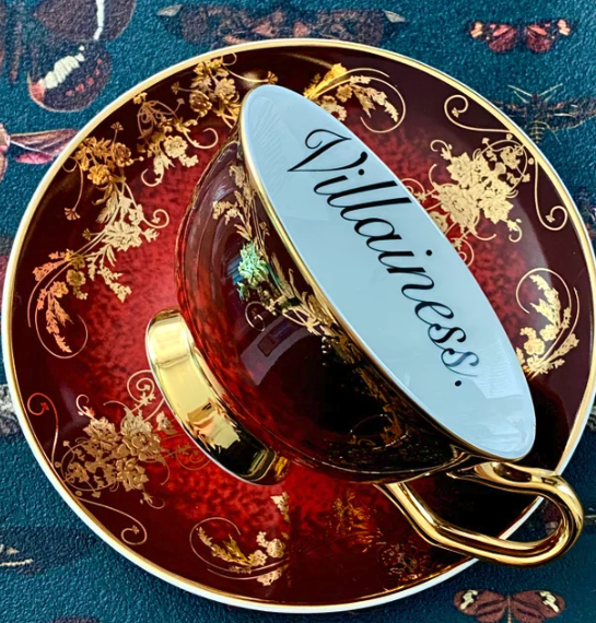 A deep red teacup and saucer with gold filigree accents and the word VILLAINESS written on the inside edge of the teacup