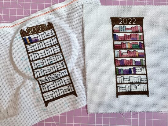Two cross stitch projects, one a 2022 complete bookshelf with little books indicating finsihed books color coded by genres - pink is romance and there's a lot of pink. The 2023 bookshelf has been started and there's a mark from the hoop frame on the aida