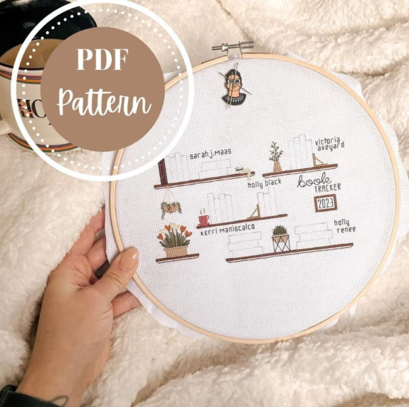A hand showing a hoop with cross stitch in progress against a furry blanket. The pattern is books on a shelf with some groups of books labeled with back stitched author names like Sarah J Maas or Victoria Aveyard