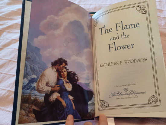 The Flame and the Flower title page with author Kathleen Woodiwiss, the Romance Collections script logo, and then a stepback image of a cliffside with giant purple clouds and two people standing in the wind with the woman in a square necked blue gown with a lot of white ruffles and a black cape billowing behind her with a man standing three quarters behind her in a giant white puffy shirt like I could live in the sleeves they are so big