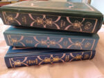 A stack of hardcover romances with blue and green colored leather an gold foil stamping stacked so the spines are visible