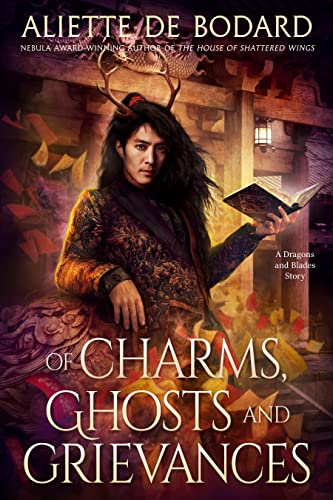 Of Charms, Ghosts, & Grievances by Aliette De Bodard. An Asian man with long hair and antlers reclines against a golden carpet. In his hand, he holds a book open.