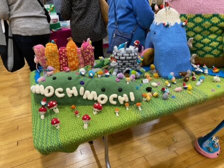 A knitted display from Mochimochi Land features a cityscape, a castle, some verdant hills and a mountain eating a donut
