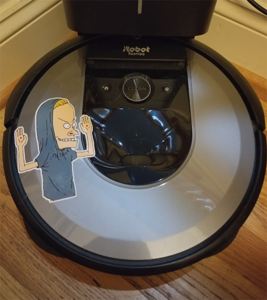 a photograph of my roomba with a beavis sticker on the left side. beavis has his shirt pulled over his head and his arms up and bent at the elbow, cornholio style