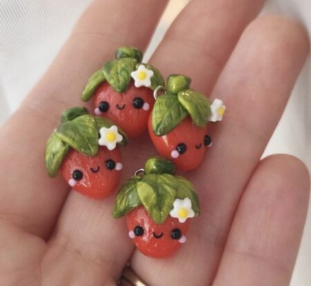 Four strawberry stitch markers held in an open palm. They have little smiley faces and flowers on their stems.