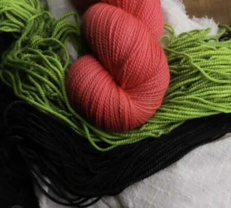 A skein of pinkish red yarn with some loose bright green and black yarn in the background.