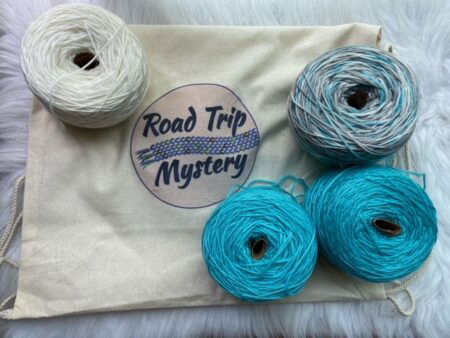 A skein of white yarn, two skeins of icy blue yarn, and a skein of blue and gray yarn on top of a bag that says Road Trip Mystery