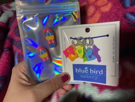 A pair of needle point protectors shaped like pink yellow and blue flip flops, and a set of neon colored stitch markers shaped like Upper Midwest states with a heart in the center of each.