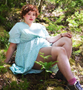 An image of a White person with red curly hair in a light blue dress showing off the shorts under their dress that are a light beige color