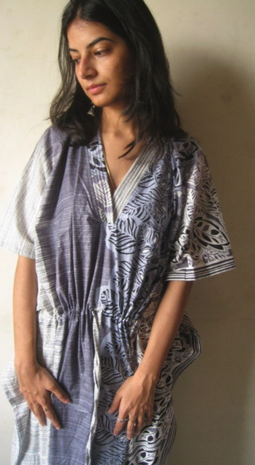 A person with shoulder length brown hair poses in a drapey soft caftan in light grey with a leaf pattern. The Caftan has a v-neck and pleats at the neck and a drawstring waist 