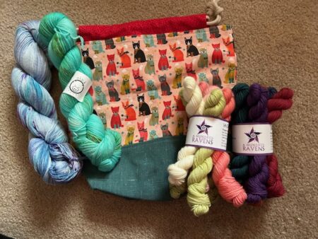 Two skeins of yarn and two mini skein sets resting against a project bag with a cat pattern.