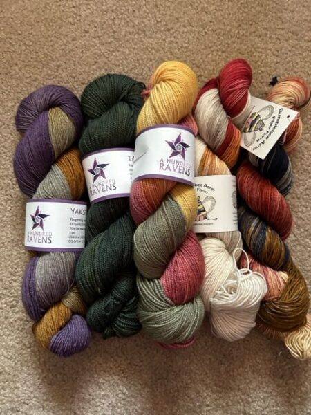 Five skeins of yarn from A Hundred Ravens in russet red, gold, and muted green with purple and grey
