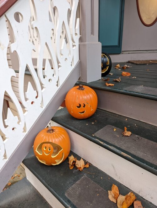 A picture of my porch steps with two orange foam pumpkins, one carved with a ghost and the other with a smiley face. There's a black pumpkin peeking from behind the railing post that has toothless from how to train your dragon carved into it. The teps are dark green and the railing has white carved panels that look like curvy lace
