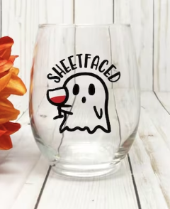 A stemless wineglass against a white wood background with a tiny ghost holding a red wine glass below the word SHEETFACED 