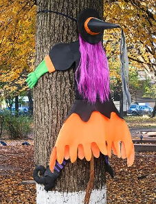A photograph of a witch with purple hair, a black hat, and orange fringed skirt and green arms hugging a tree like it just flew straight into it 
